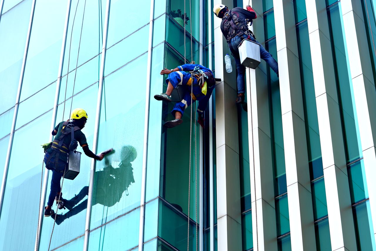 why-choose-us Three window cleaners hanging from ropes clean the facade of a modern glass building in daylight.
